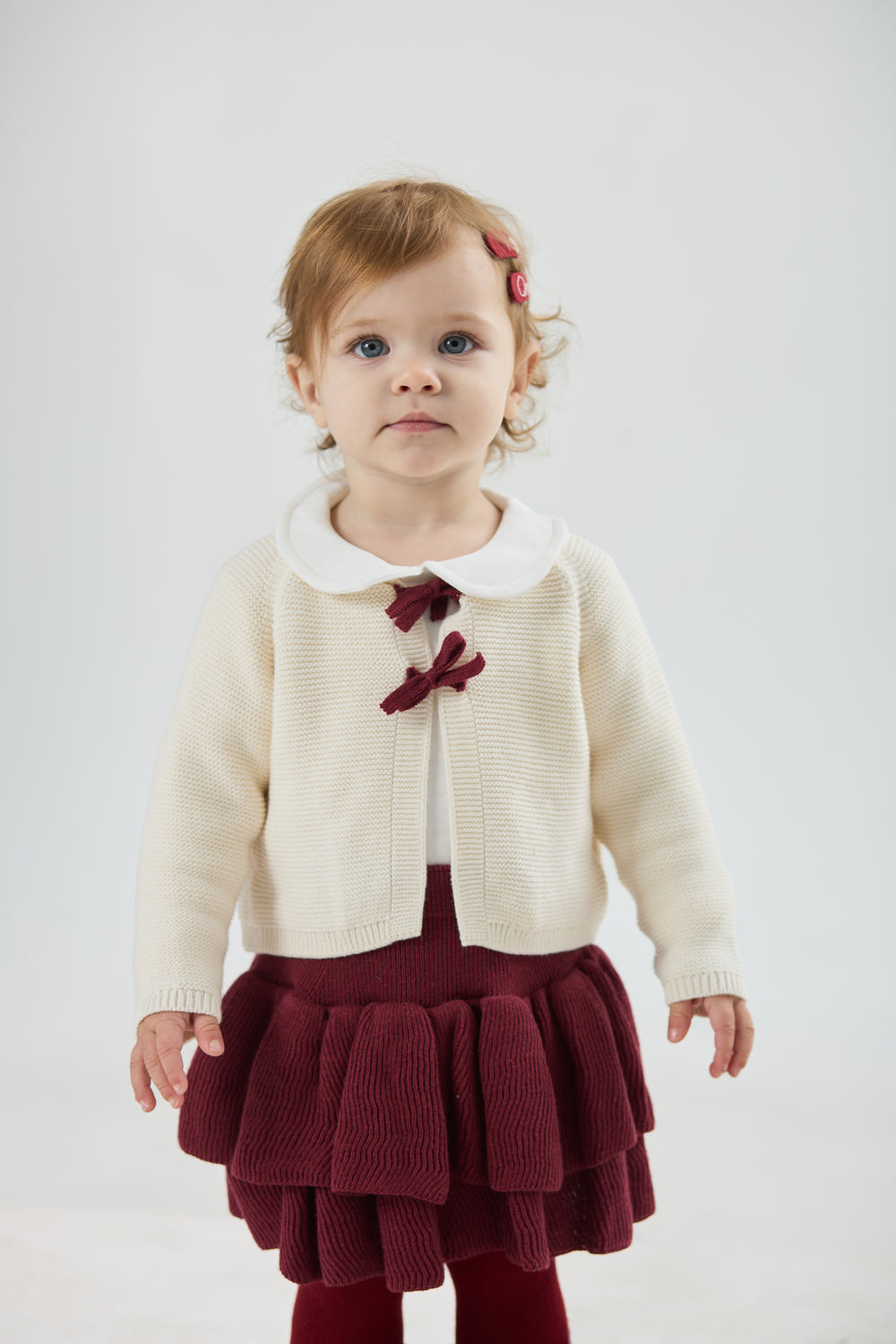 A young girl holding out the skirt of a deep burgundy velvet dress trimmed with white lace